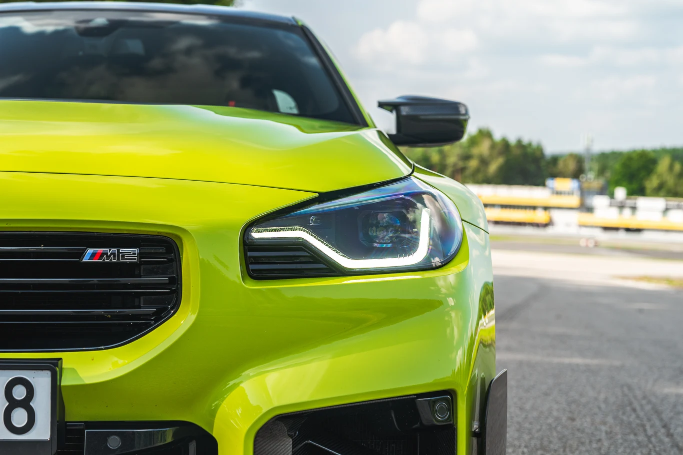 BMW M2 Jaskrawożółty sportowy samochód BMW M2 z charakterystycznym światłem LED, ustawiony na torze wyścigowym, w tle widoczne fragmenty toru i barierki oraz rozmyta zieleń.