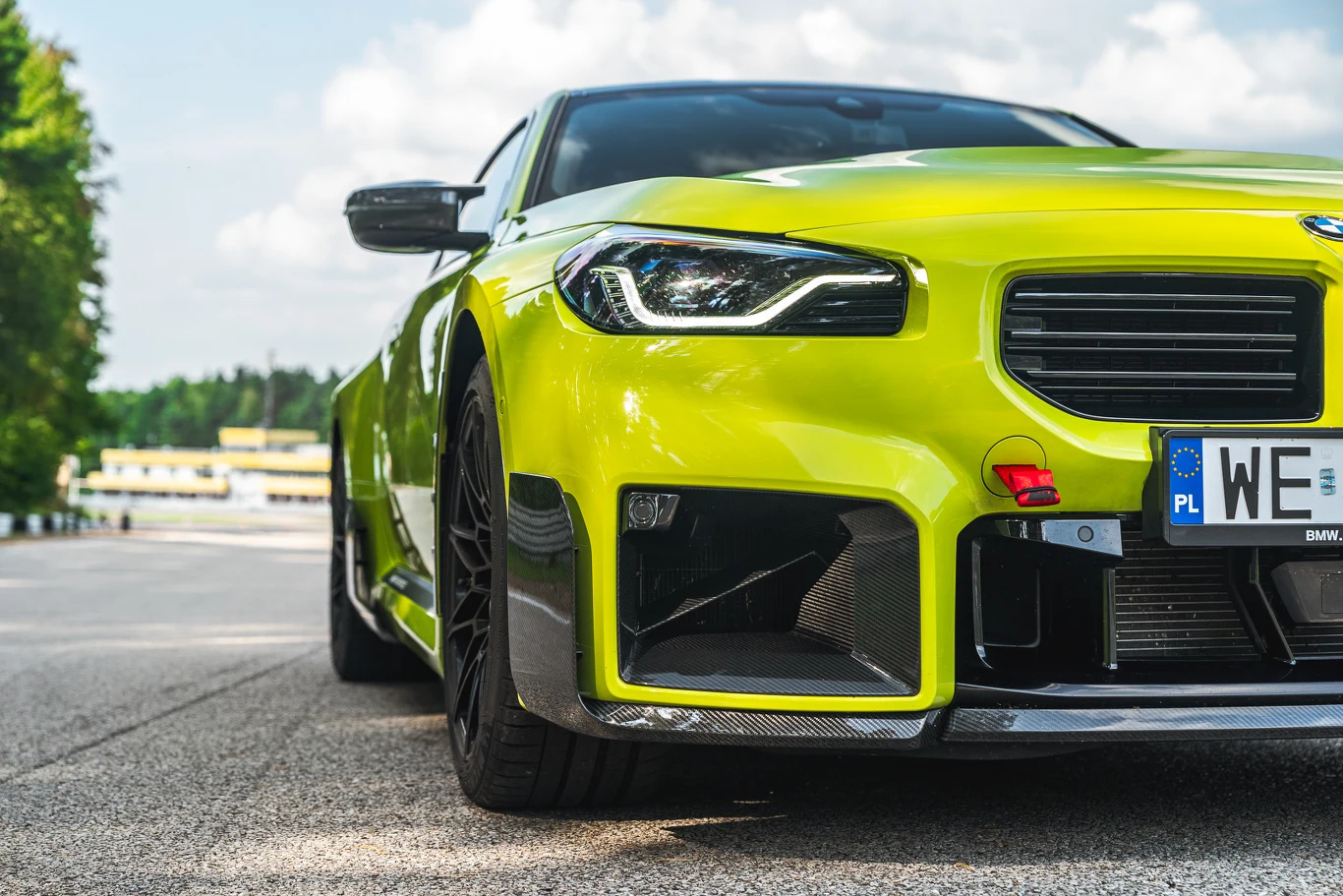 BMW M2 Jaskrawozielony sportowy samochód BMW ustawiony w lewym dolnym rogu kadru, widok na przód pojazdu z detalami zderzaka, reflektorów i tablicy rejestracyjnej, w tle tor wyścigowy i drzewa po lewej stronie.