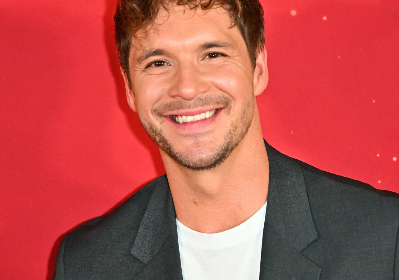 Mikołaj Roznerski Uśmiechnięty mężczyzna z krótkimi, brązowymi włosami i lekkim zarostem w ciemnej marynarce i białym T-shircie na jednolitym czerwonym tle.