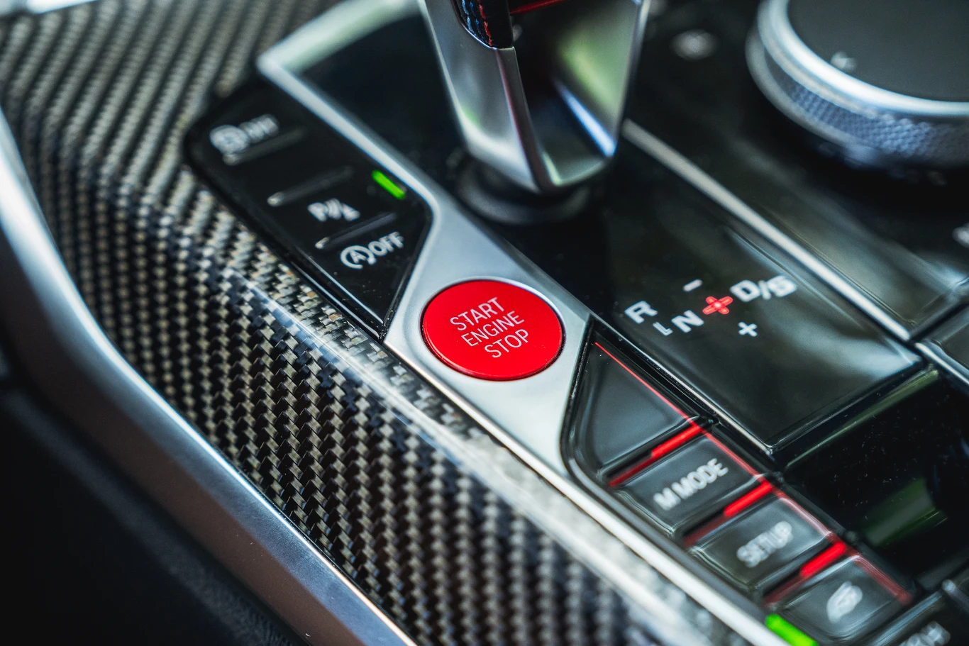 BMW M2 Fragment nowoczesnej konsoli środkowej samochodu z karbonowym wykończeniem, przyciskiem Start Engine Stop w kolorze czerwonym, dźwignią zmiany biegów oraz przyciskami do sterowania systemem jazdy.