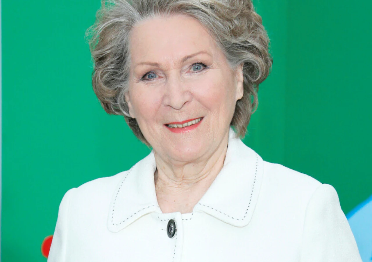 Irena Santor Starsza kobieta z siwymi, delikatnie ułożonymi włosami stoi na tle zielonej powierzchni, ubrana w elegancki biały żakiet i delikatnie się uśmiecha, patrząc prosto w obiektyw.