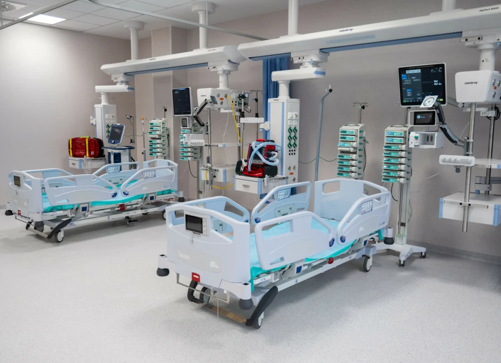 Dwa nowoczesne łóżka szpitalne otoczone zaawansowanym sprzętem medycznym, monitorami oraz urządzeniami do podtrzymywania życia, ustawione w jasnym, czystym pomieszczeniu szpitalnym.