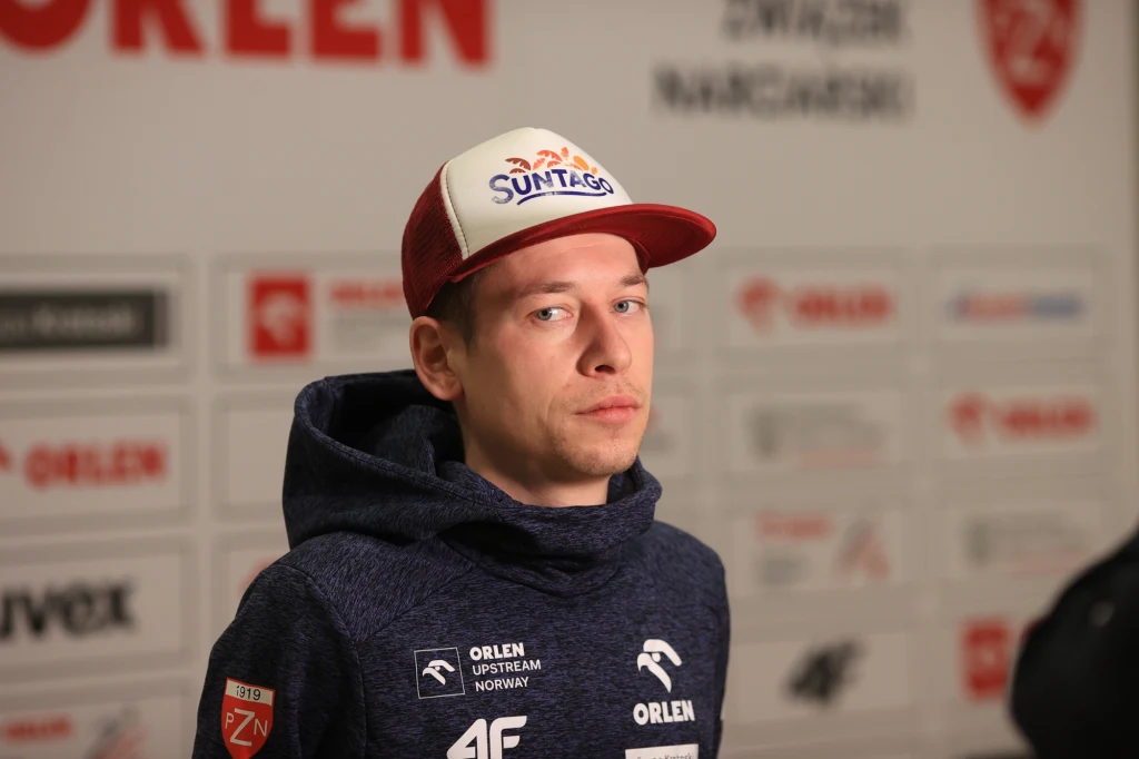 Aleksander Zniszczoł Mężczyzna w sportowej bluzie i czapce z daszkiem z logotypami sponsorów na tle ściany z logotypami partnerów oraz informacji prasowych, patrzy w stronę obiektywu z poważnym wyrazem twarzy.