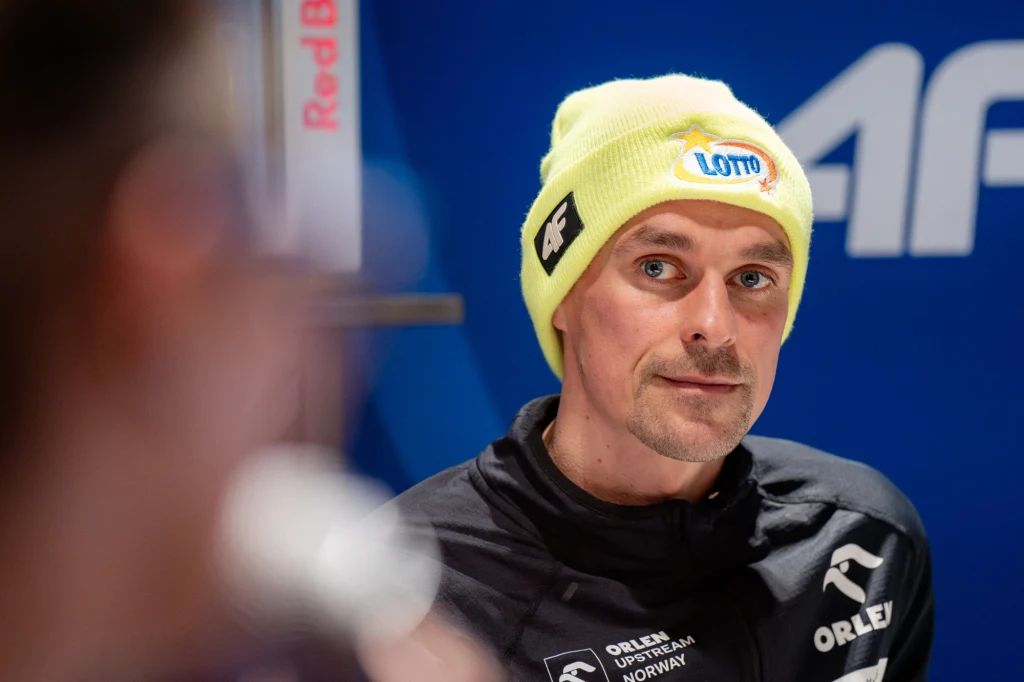 Piotr Żyła szuka spokoju w Pucharze Świata Mężczyzna w jasnej żółtej czapce z logo Lotto oraz czarnej sportowej kurtce z widocznymi logotypami sponsorów, patrzący spokojnie w stronę kamery, na tle granatowej ścianki z elementami brandingowymi.