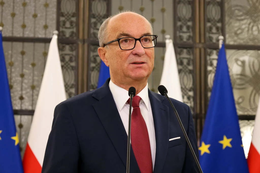 Mężczyzna w garniturze i czerwonym krawacie stoi przed mikrofonami na tle flag Polski i Unii Europejskiej.