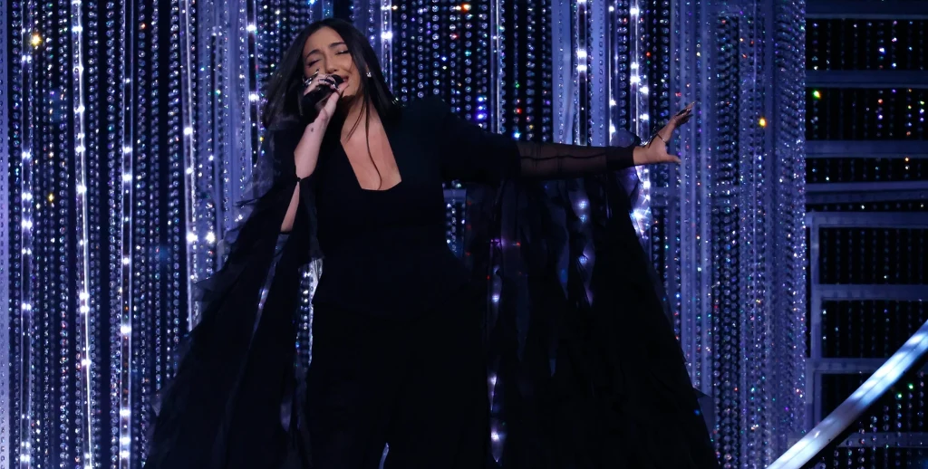 Izrael wystąpi podczas Eurowizji 2026 Kobieta ubrana na czarno śpiewa na scenie, trzyma mikrofon w prawej dłoni, za jej plecami widoczne są błyszczące, dekoracyjne kurtyny oraz fragment schodów.