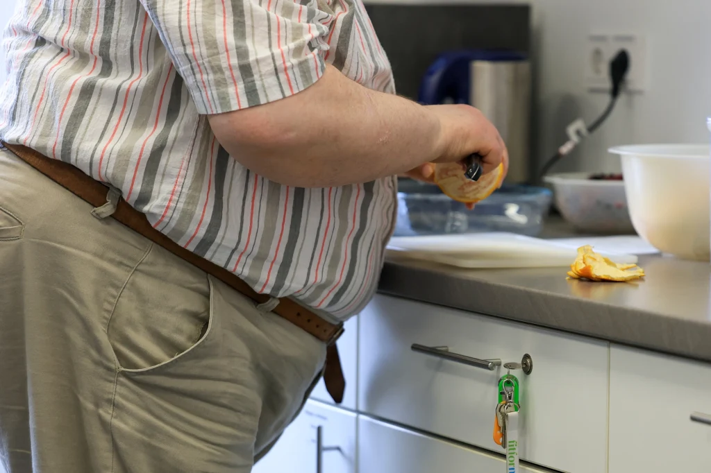 Średnia długość życia w Niemczech spada, a ma to związek m.in. z nieprawidłowym odżywianiem - informuje "Deutsche Welle" Osoba otyła kroi owoc na blacie kuchennym w pomieszczeniu kuchennym. Widoczne są blat z plastikową miską, owoce oraz fragmenty przedmiotów kuchennych.