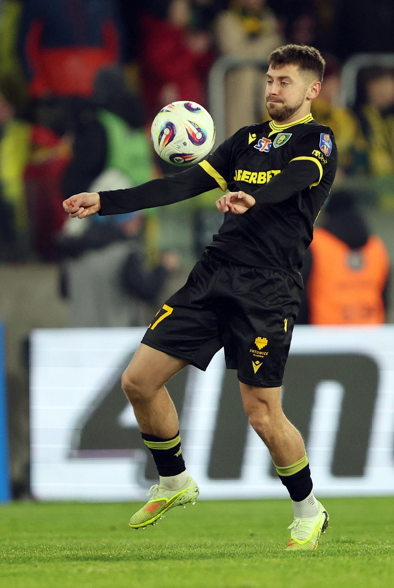 Piłkarz w czarno-żółtym stroju wykonuje precyzyjne zagranie piłką na stadionie, w tle zamazana publiczność i jasno oświetlony baner reklamowy.
