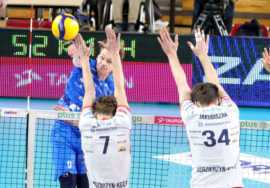 Intensywna akcja podczas meczu siatkówki, zawodnik w niebieskim stroju wykonuje atak, a dwóch graczy drużyny przeciwnej próbuje zablokować piłkę pod siatką.