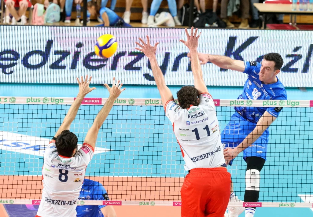 Zawodnik w niebieskim stroju atakuje piłkę podczas meczu siatkówki, próbując przebić ją przez blok dwóch przeciwników w białych koszulkach i czerwonych spodenkach, na tle widoczna siatka oraz fragmenty trybun.