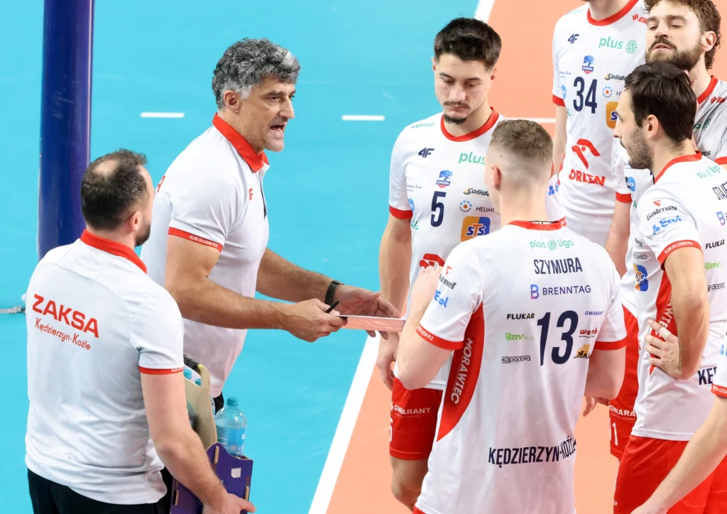 Trener drużyny siatkarskiej udzielający wskazówek zawodnikom podczas przerwy w meczu, wszyscy ubrani w oficjalne stroje klubowe z widocznymi nazwiskami i numerami.