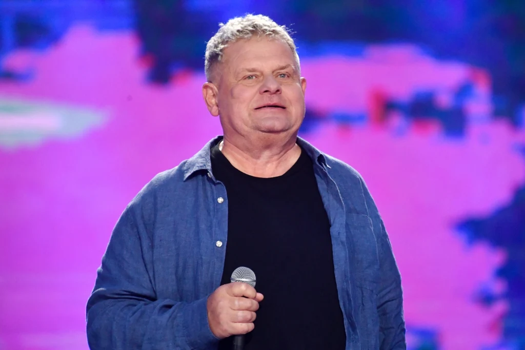 Mężczyzna trzymający mikrofon, ubrany w ciemną koszulkę i niebieską koszulę, stoi na tle kolorowego, rozmytego ekranu.