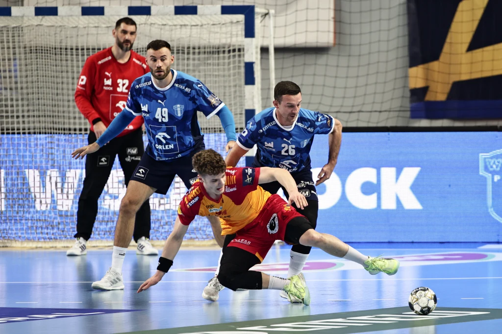 Zawodnicy grający w piłkę ręczną podczas intensywnej akcji na parkiecie, jeden z zawodników w żółto-czerwonym stroju upada na ziemię, rywale w niebieskich koszulkach próbują przejąć piłkę, w tle widoczny bramkarz i bramka.