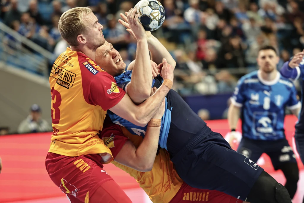 Dynamiczna akcja podczas meczu piłki ręcznej, zawodnik w niebieskim stroju próbuje rzucić piłkę do bramki, będąc intensywnie blokowany przez dwóch rywali w żółto-czerwonych strojach. W tle widoczne trybuny pełne kibiców oraz pozostali zawodnicy.