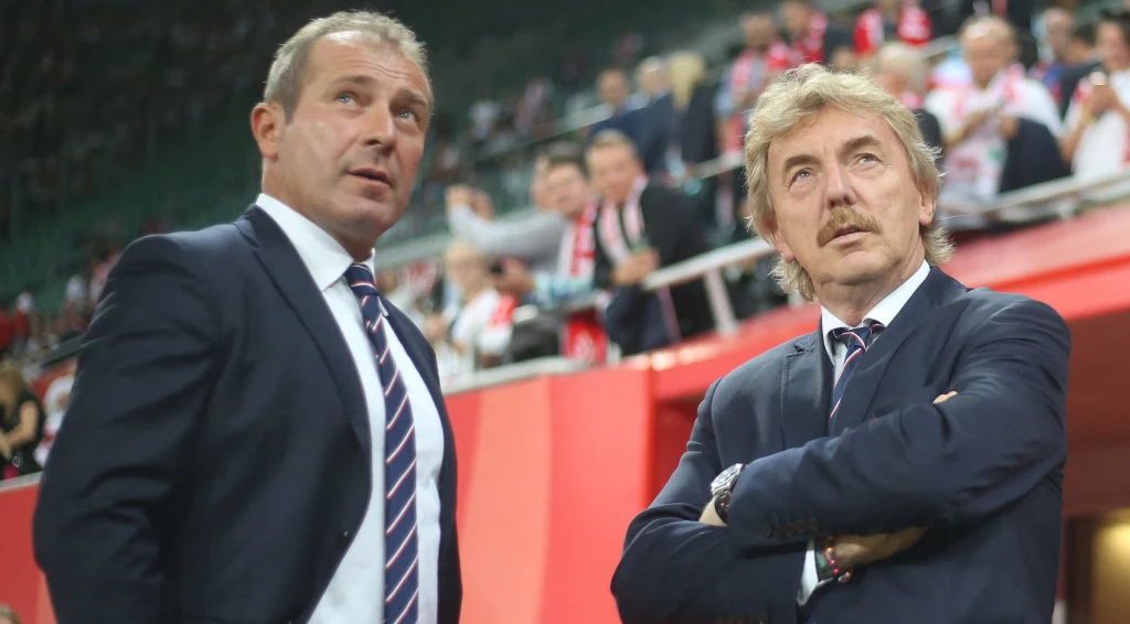 Dwóch mężczyzn w garniturach stoi na tle trybun stadionu, w tle widoczna grupa ludzi w biało-czerwonych barwach. Obaj panowie wyglądają na skupionych, jeden z nich ma skrzyżowane ręce.