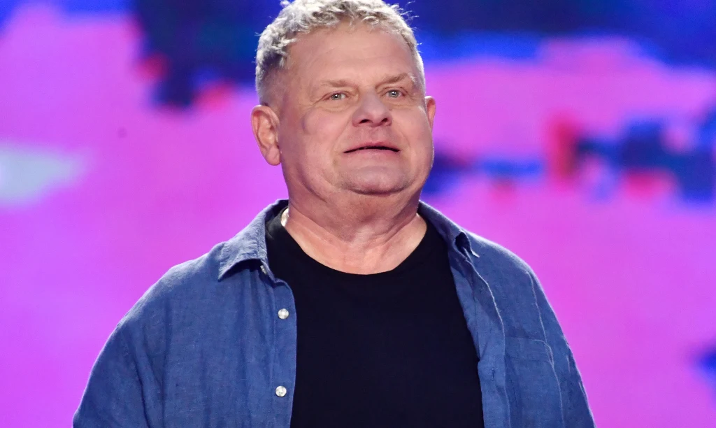 Kazik Staszewski Doświadczony mężczyzna z siwymi włosami w niebieskiej koszuli i czarnym podkoszulku stoi na tle rozmytego, kolorowego tła.