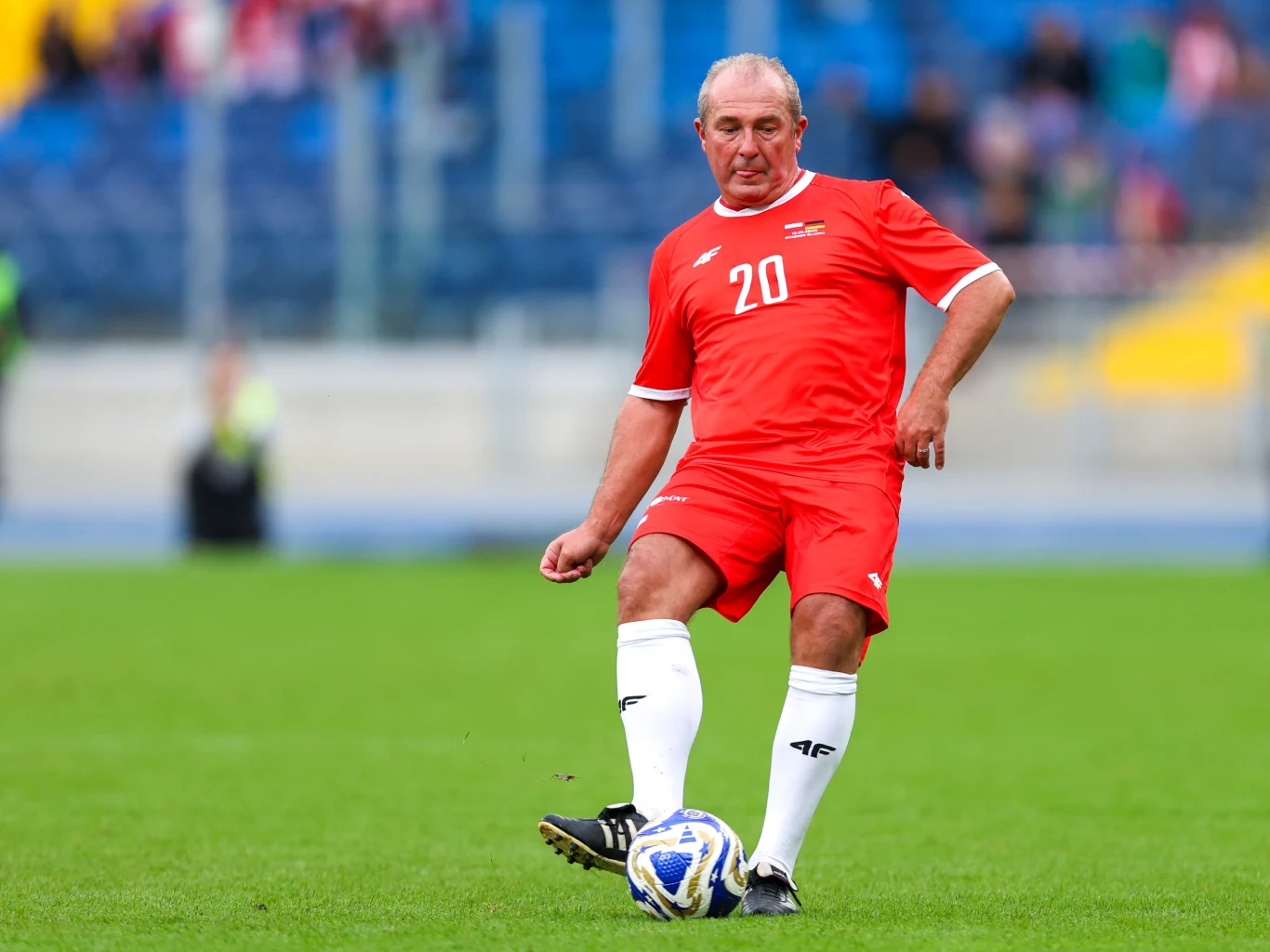 Starszy mężczyzna ubrany w czerwony strój piłkarski z numerem 20 na boisku podczas gry w piłkę nożną, koncentrujący się na prowadzeniu piłki.