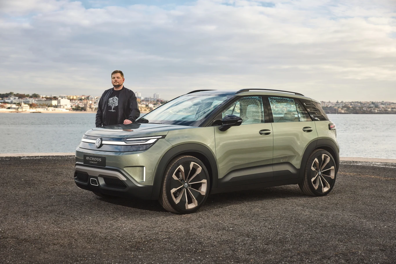 Nowoczesny samochód elektryczny w jasnoszarym kolorze ustawiony na tle wody i panoramy miasta, obok samochodu stoi mężczyzna w casualowym ubraniu.