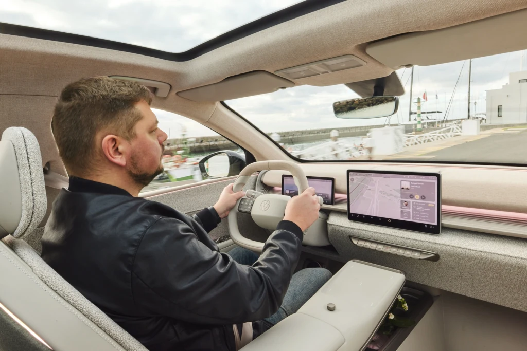 Mężczyzna prowadzi nowoczesny samochód z panoramicznym dachem i zaawansowanym systemem multimedialnym widocznym na dużym ekranie dotykowym przy desce rozdzielczej. Jasne wnętrze pojazdu oraz widok na marinę przez okno podkreślają komfortową atmosferę.