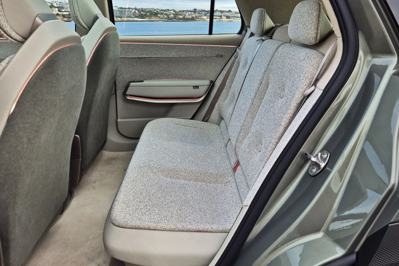 Tylny rząd siedzeń samochodu osobowego z jasnoszarymi tapicerkami, widok na siedzenia i drzwi, w tle przez okno widoczny fragment krajobrazu z wodą i zabudowaniami.