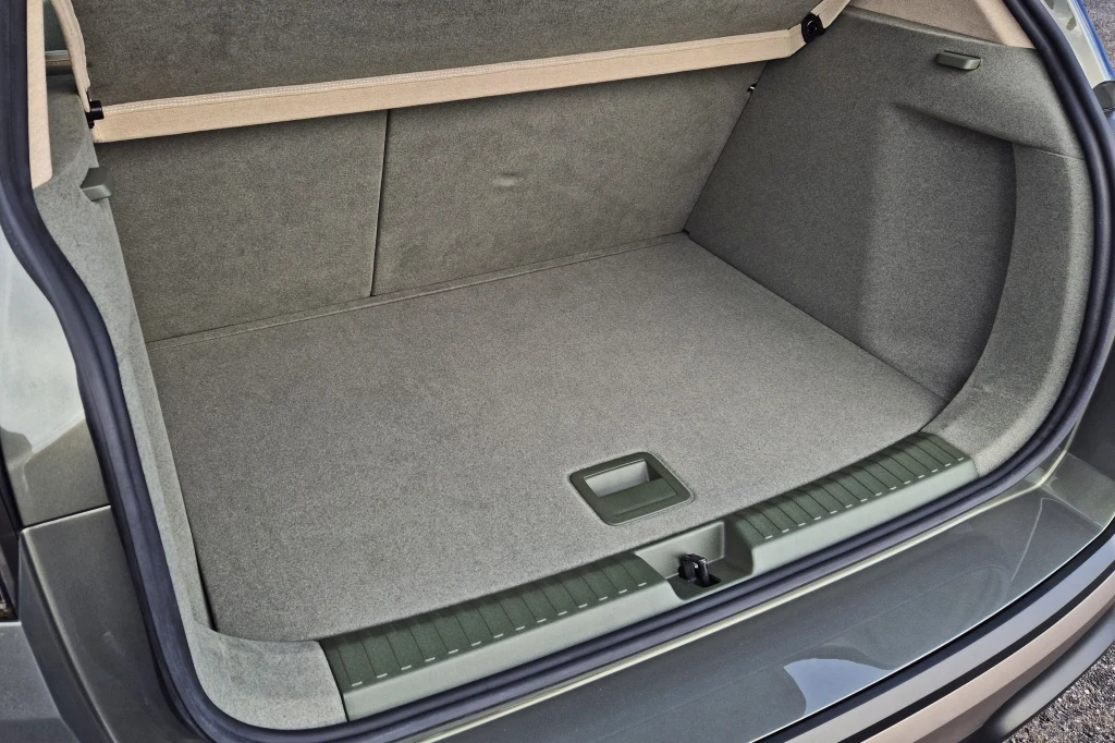 Pojemny bagażnik samochodowy wyłożony szarym materiałem, zamknięcie z tworzywa sztucznego pośrodku podłogi, krawędź zabezpieczona plastikowym elementem, otwarte drzwi bagażnika.