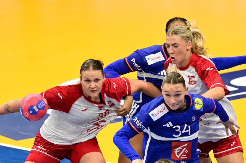 Dynamiczna walka zawodniczek podczas meczu piłki ręcznej, zawodniczki w białych i czerwonych strojach próbują zatrzymać przeciwniczkę w niebieskim stroju na żółtym parkiecie.