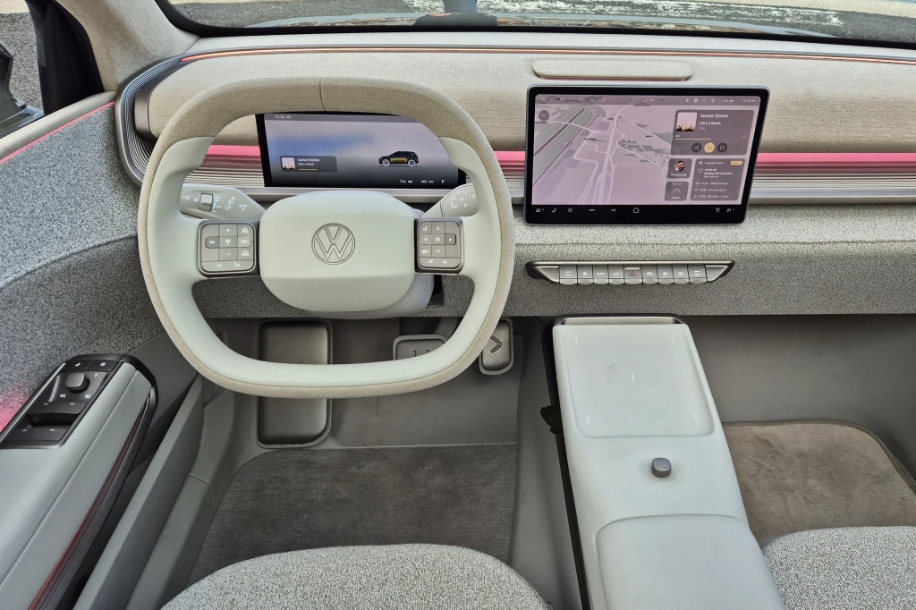 Kokpit nowoczesnego samochodu marki Volkswagen z jasnym, minimalistycznym wykończeniem. Centralnie widoczna kierownica z futurystycznym projektem, dwa duże ekrany cyfrowe, w tym jeden prezentujący mapę nawigacji i cyfrowe sterowanie pojazdu. Panel ster...