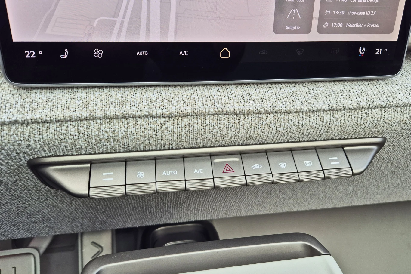 Panel klimatyzacji samochodowej z przyciskami sterującymi nawiewem, klimatyzacją, trybem automatycznym, podgrzewaniem oraz światłami awaryjnymi; powyżej widoczny fragment ekranu dotykowego z cyfrowym interfejsem systemu multimedialnego.