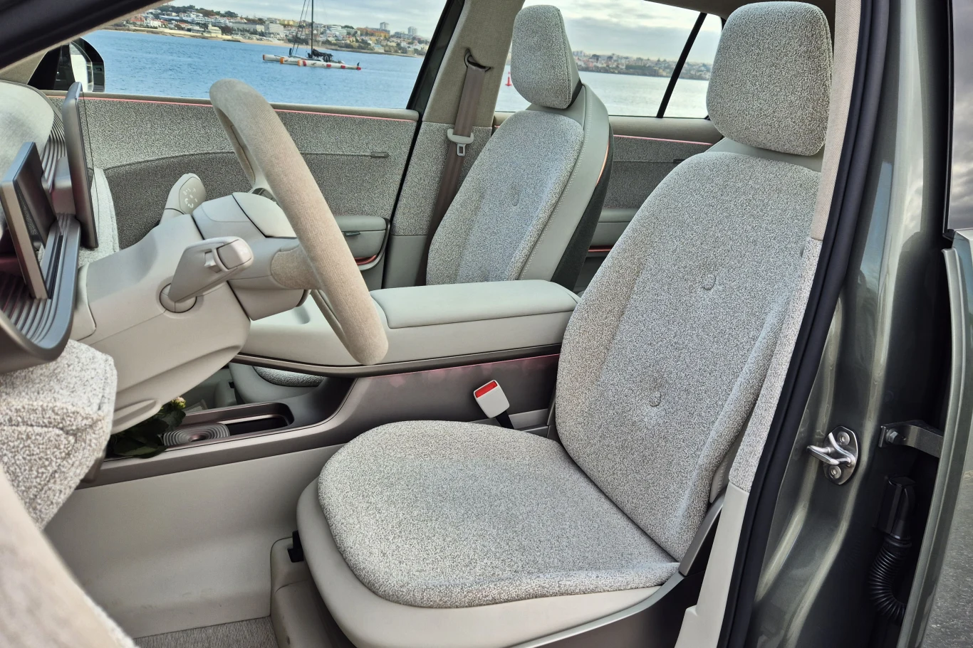 Wnętrze nowoczesnego samochodu z jasnoszarymi fotelami obitymi materiałem oraz minimalistyczną deską rozdzielczą, przez okna widoczny krajobraz z wodą i zabudowaniami w oddali.