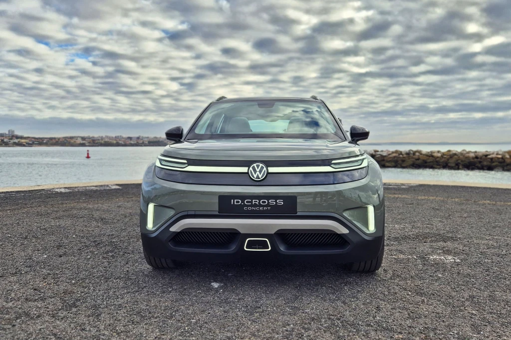 Samochód elektryczny Volkswagen ID. Cross ustawiony na asfaltowej nawierzchni z widokiem na wodę i kamienisty brzeg w tle, pod pochmurnym niebem.