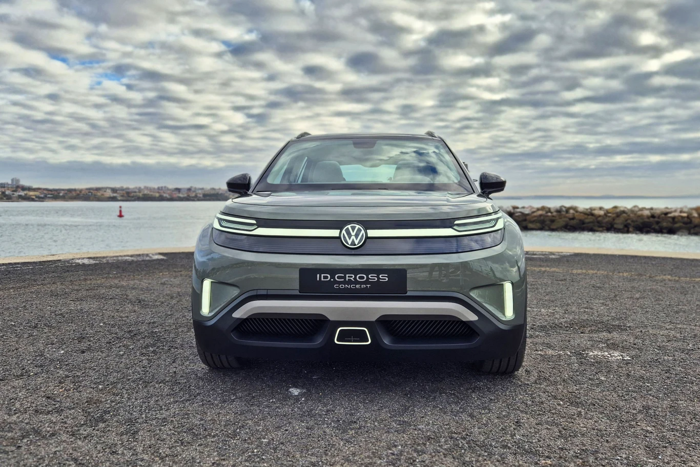 Samochód elektryczny Volkswagen ID. Cross ustawiony na asfaltowej nawierzchni z widokiem na wodę i kamienisty brzeg w tle, pod pochmurnym niebem.