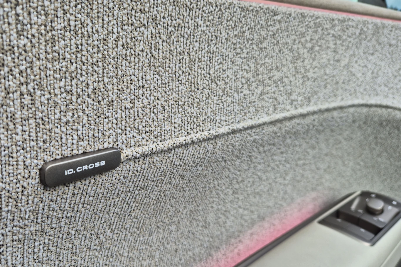 Fragment drzwi samochodu z tekstylną tapicerką w jasnoszarym kolorze, oznaczeniem 'ID.CROSS' oraz widocznym panelem sterowania szybą.