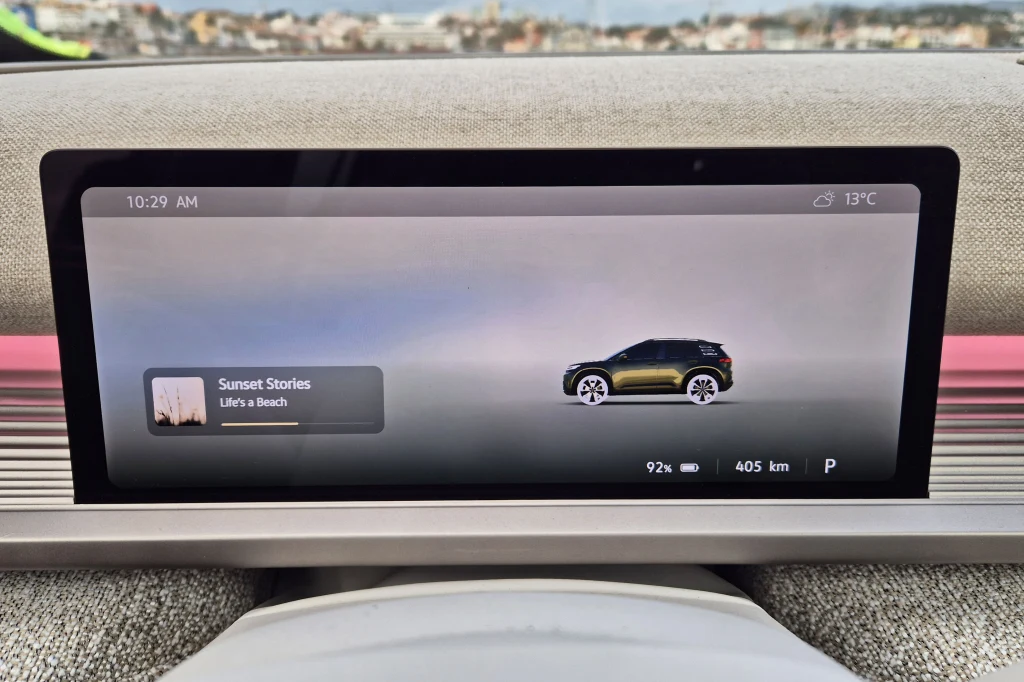 Wnętrze samochodu z nowoczesnym, szerokim ekranem dotykowym na środku deski rozdzielczej. Na ekranie widoczna jest grafika samochodu, poziom naładowania baterii, zasięg oraz temperatura zewnętrzna. W tle przez szybę można dostrzec rozmyte budynki i poc...