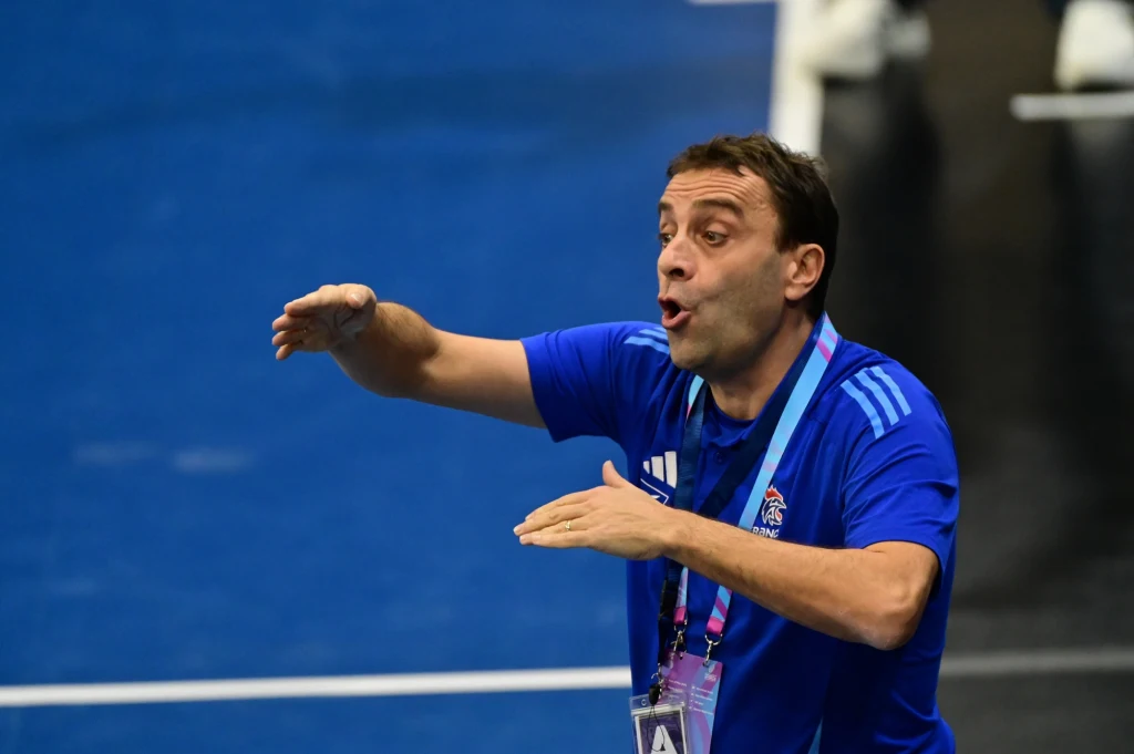 Mężczyzna w niebieskiej koszulce sportowej gestykuluje energicznie, stojąc na hali sportowej. Na jego szyi zawieszony jest identyfikator, a w tle widoczna jest nieostra część boiska.