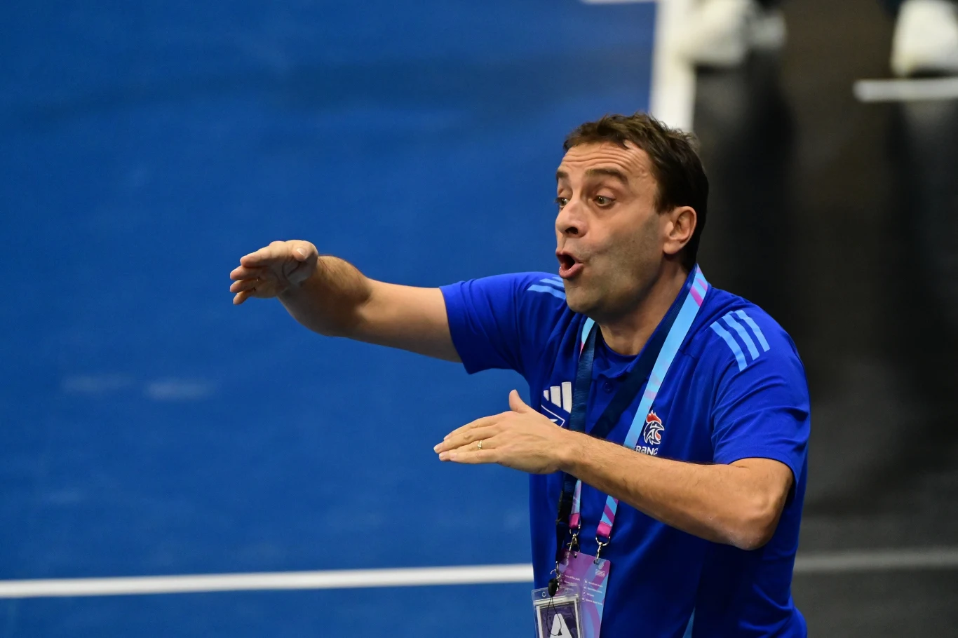 Mężczyzna w niebieskiej koszulce sportowej gestykuluje energicznie, stojąc na hali sportowej. Na jego szyi zawieszony jest identyfikator, a w tle widoczna jest nieostra część boiska.