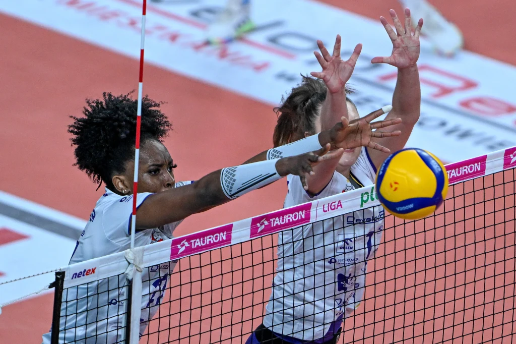 Blok siatkarek DevelopResu Rzeszów Dwie zawodniczki siatkarskie w trakcie dynamicznej akcji przy siatce próbujące zablokować przeciwnika, żółto-niebieska piłka siatkowa widoczna nad siatką, fragment boiska i siatki ozdobionej logotypami sponsorów.