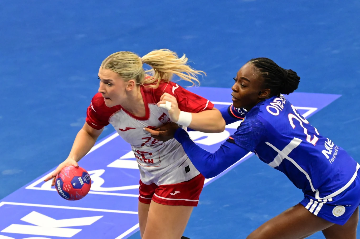 Dwie zawodniczki podczas meczu piłki ręcznej, jedna ubrana w biało-czerwony strój próbuje przedostać się z piłką przez obronę przeciwniczki w niebieskim stroju, na tle niebieskiego boiska.