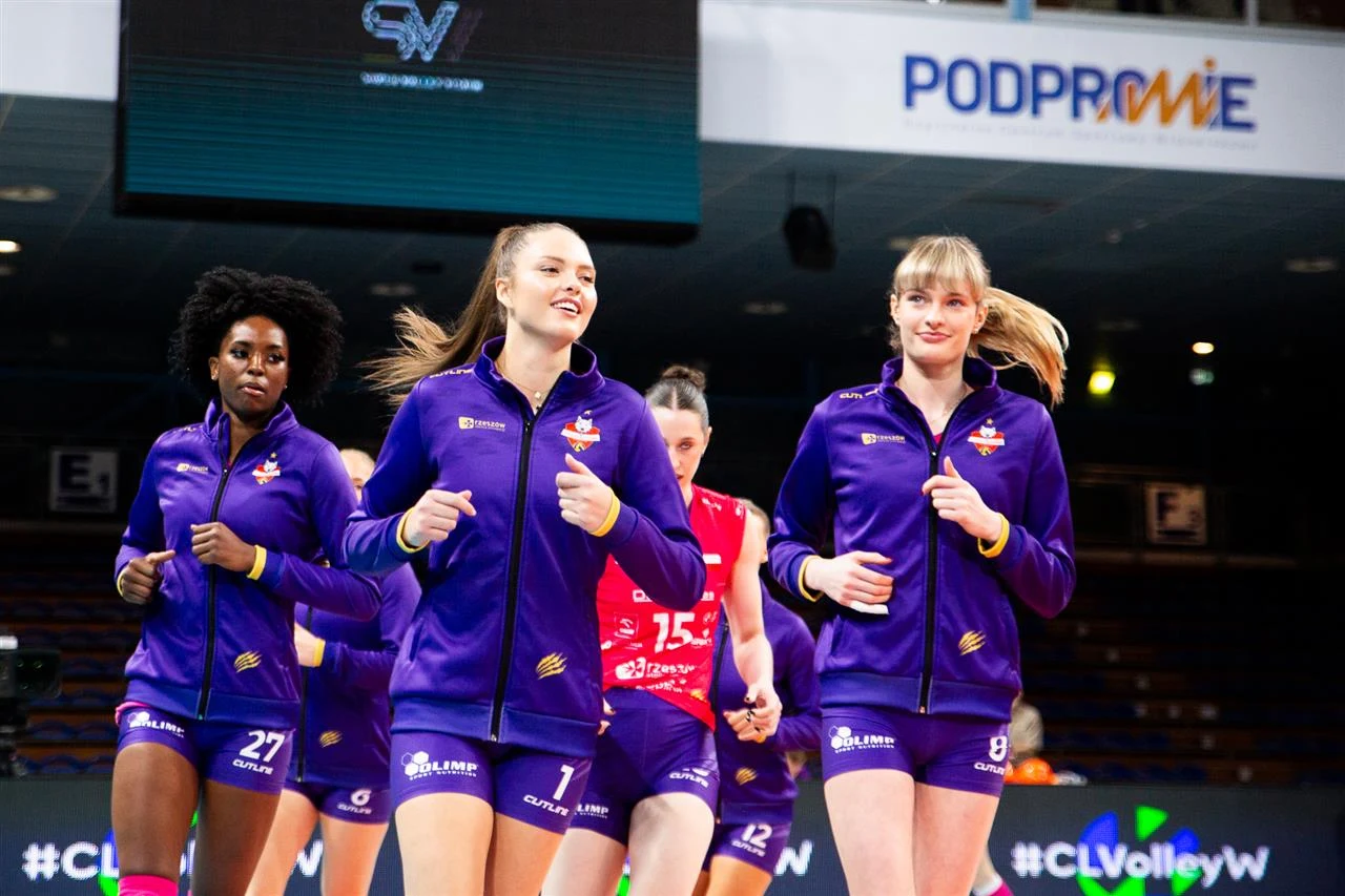 Siatkarki w fioletowych dresach wykonujące rozgrzewkę na hali sportowej, w tle widoczne trybuny oraz logotypy sponsorów nad boiskiem.
