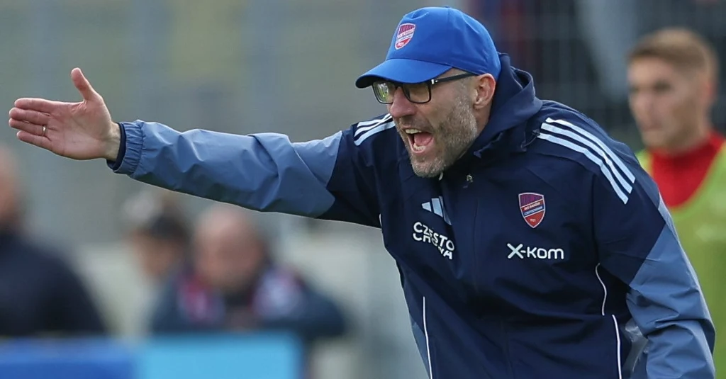 Marek Papszun, trener Rakowa Częstochowa Trener drużyny piłkarskiej w niebieskiej czapce i kurtce energicznie gestykuluje podczas meczu, pokazując zaangażowanie. W tle niewyraźne postacie innych członków zespołu oraz trybuny.