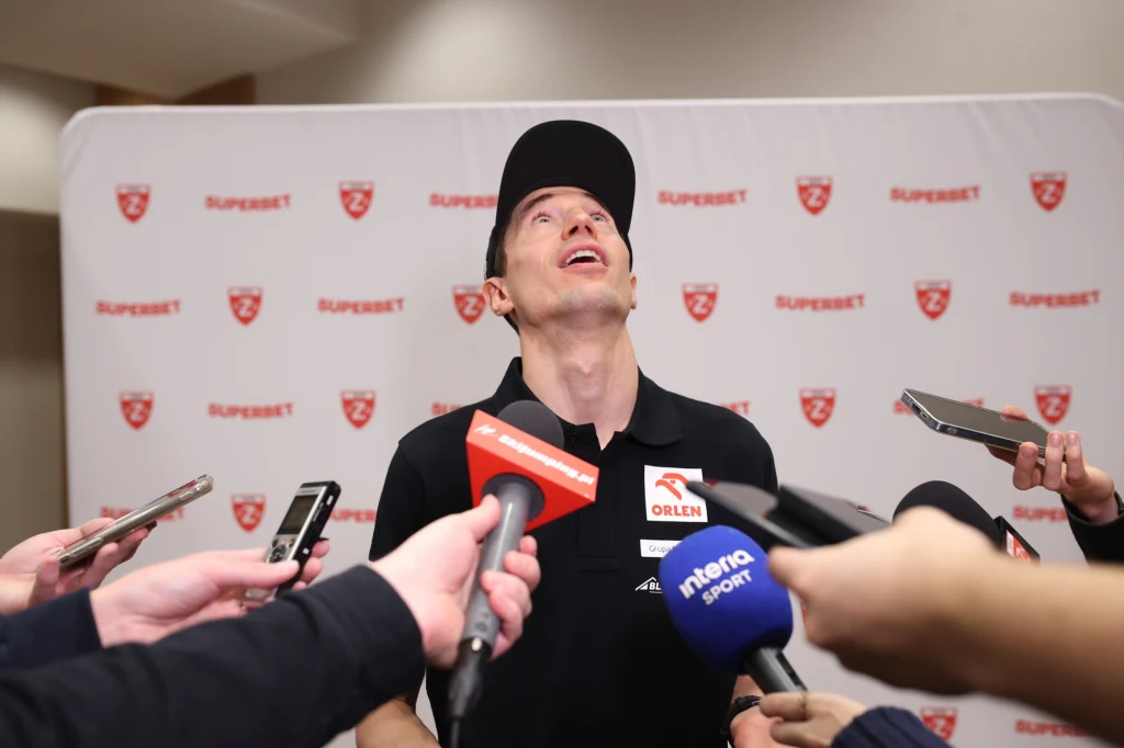 Mężczyzna w czarnej koszulce oraz czapce z daszkiem stoi przed ścianką z logotypami i patrzy w górę, wokół niego trzymane są mikrofony oraz dyktafony przez dziennikarzy.