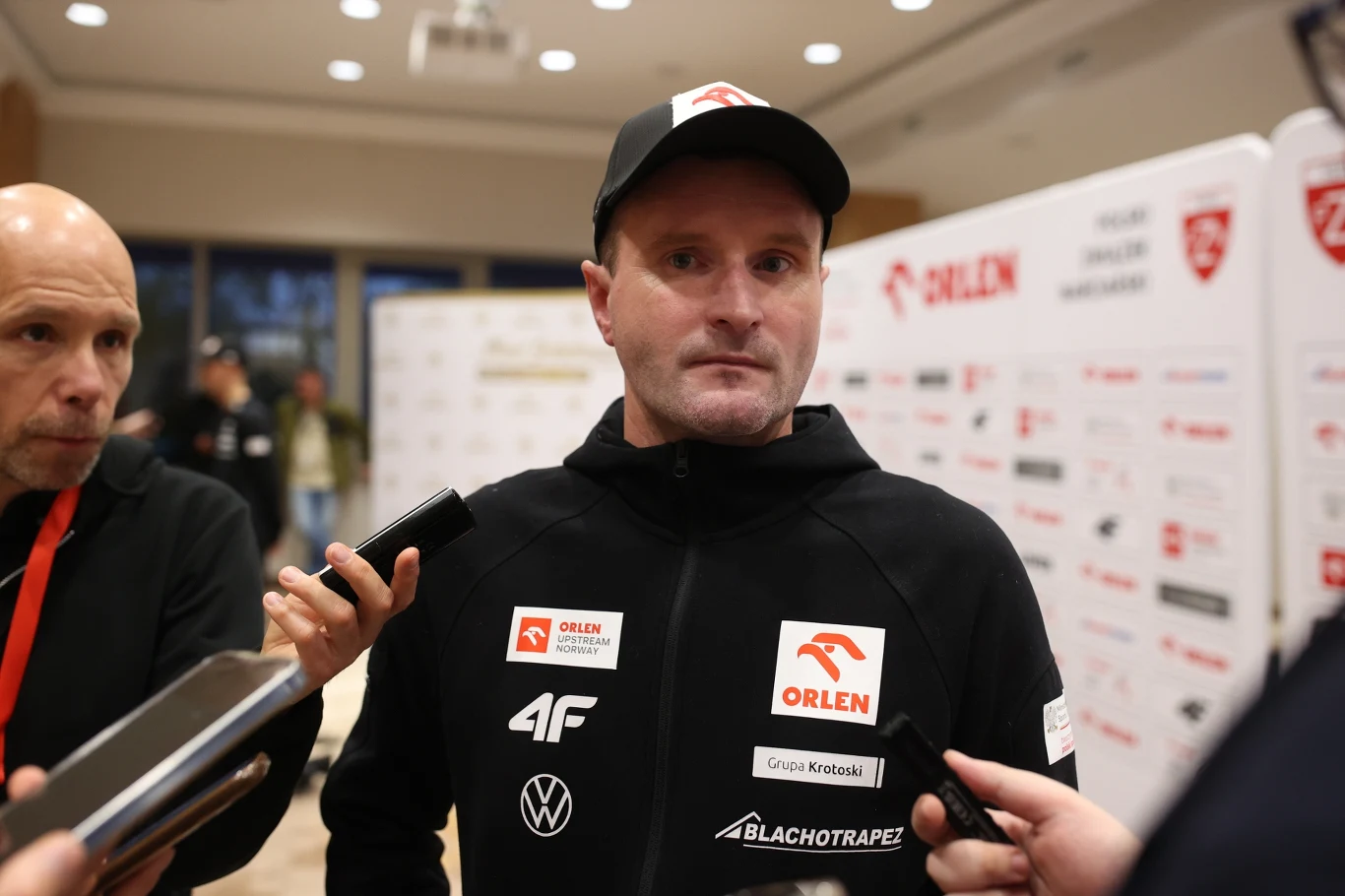 Maciej Maciusiak Mężczyzna ubrany w czarną sportową bluzę i czapkę z logotypami sponsorów udziela wywiadu kilku dziennikarzom, przy jednym ze stoisk medialnych na tle tablicy z logo Orlen i innymi oznaczeniami partnerów.