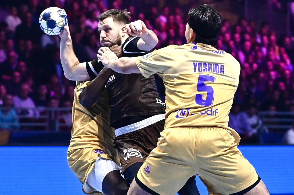 Aleks Vlah przedziera się przez defensywę Nantes Dwóch zawodników w żółtych strojach próbuje powstrzymać atakującego szczypiornistę w czarnym stroju, który trzyma piłkę w wyciągniętej ręce. W tle widać publiczność oraz oświetlone trybuny.