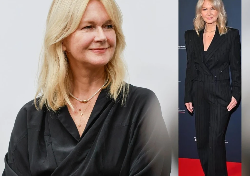 Blondynka w średnim wieku ubrana w czarny elegancki garnitur, uśmiechnięta, nosi delikatny naszyjnik. Po lewej stronie widoczny portret kobiety, po prawej pełna sylwetka na czerwonym dywanie na tle ściany z napisami.