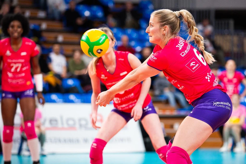 Przyjmująca DevelopResu Rzeszów Marrit Jasper Zawodniczka w różowym stroju odbija piłkę siatkową, a w tle widać koleżanki z drużyny i obserwującą widownię. Wszystkie zawodniczki skupione na przebiegu gry.