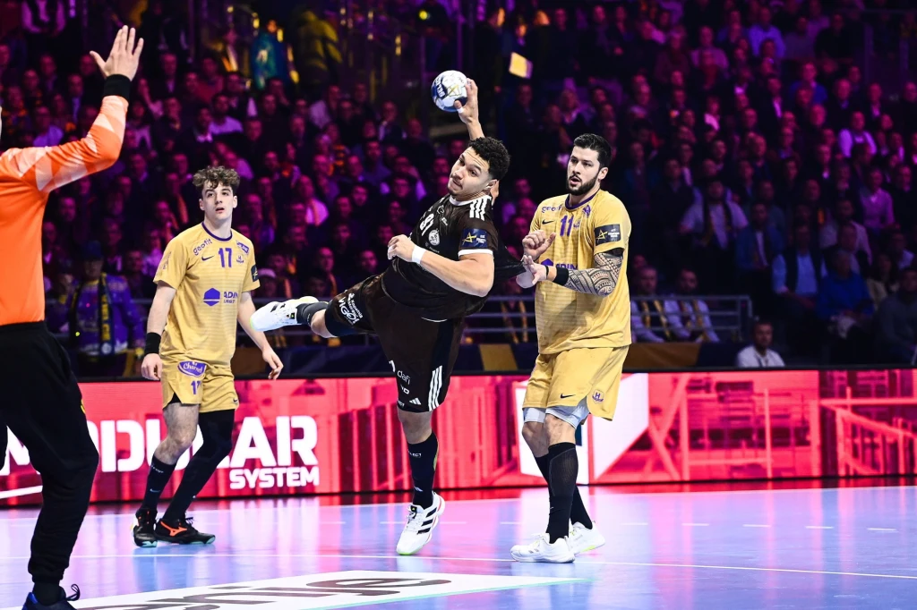 Theo Monar w meczu HBC Nantes - Industria Kielce Zawodnik w czarnym stroju wykonuje rzut w stronę bramki podczas meczu piłki ręcznej, otoczony przez dwóch obrońców w żółtych strojach, na pierwszym planie widoczny również bramkarz i sędzia, w tle publiczność.