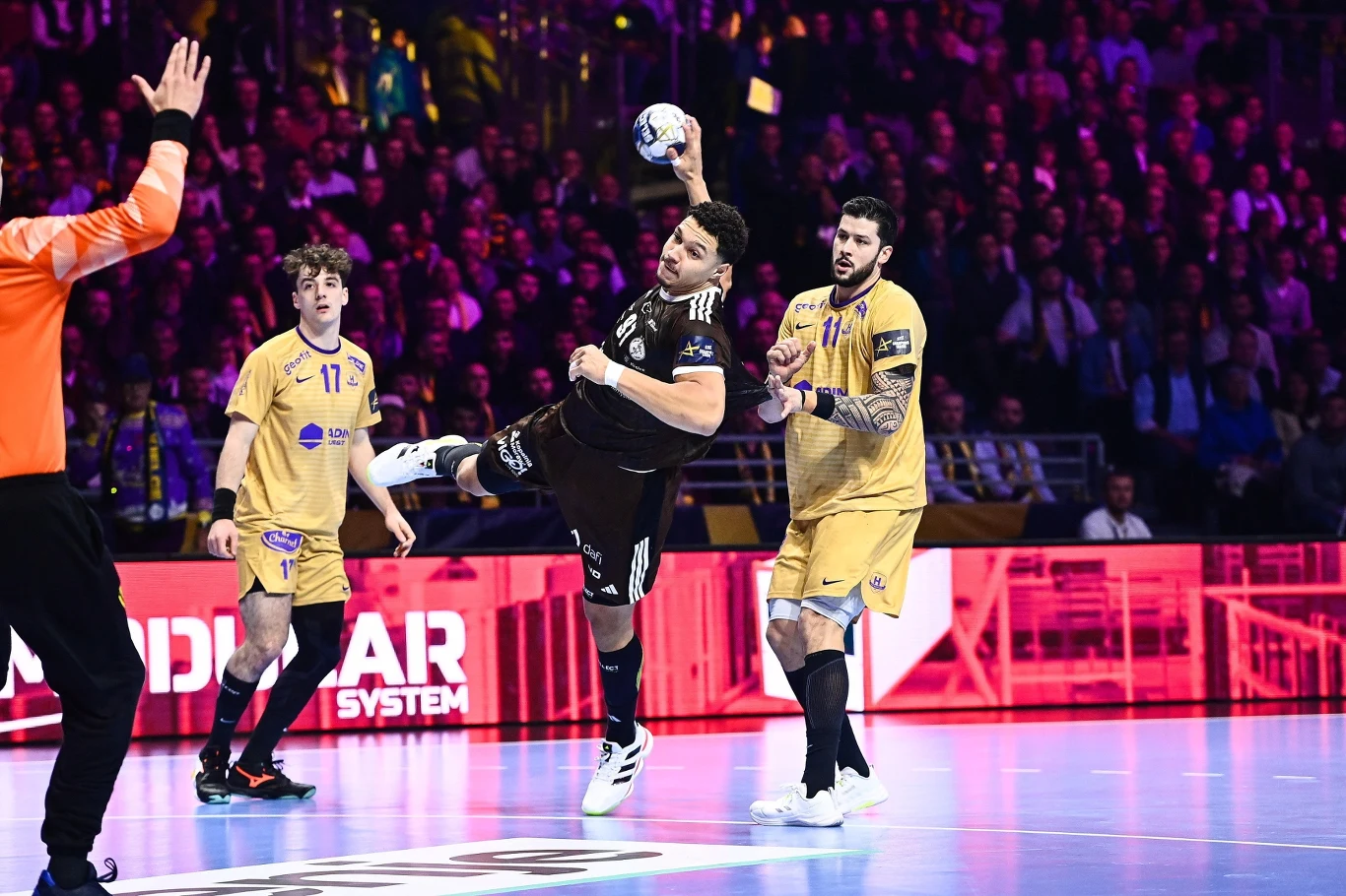 Zawodnik w czarnym stroju wykonuje rzut w stronę bramki podczas meczu piłki ręcznej, otoczony przez dwóch obrońców w żółtych strojach, na pierwszym planie widoczny również bramkarz i sędzia, w tle publiczność.