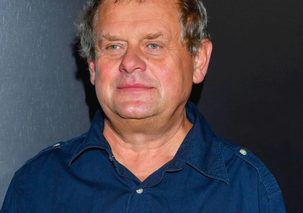 Kazik Staszewski Starszy mężczyzna w granatowej koszuli na tle ciemnej ściany, wyraz twarzy spokojny, włosy siwe, lekko uśmiechnięty.