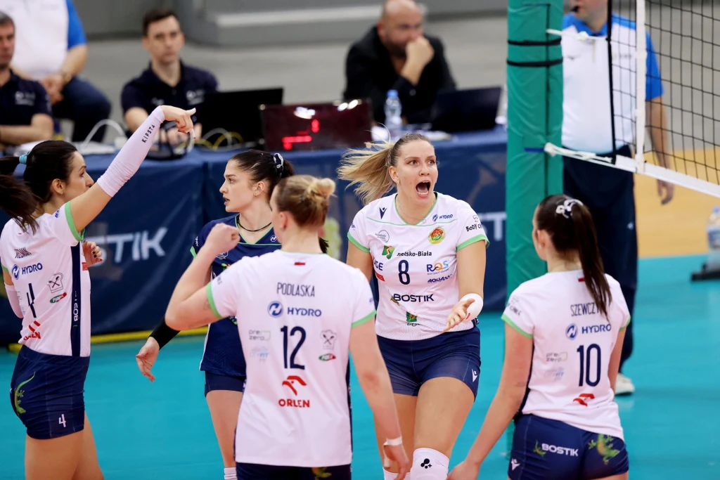 Siatkarki z Bielska-Białej Zawodniczki drużyny siatkarskiej w białych i niebieskich strojach świętują zdobycie punktu na boisku podczas meczu, jedna z zawodniczek jest wyraźnie podekscytowana i unosi rękę, w tle widać siatkę oraz sztab trenerski przy stoliku.