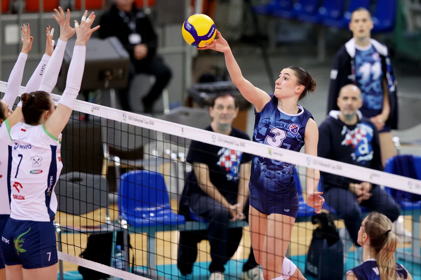 Siatkarki z Bielska-Białej zagrają w kolejnej rundzie Pucharu CEV Siatkarka drużyny w granatowych strojach przebija piłkę przez siatkę, atakując podczas meczu, naprzeciw niej dwie zawodniczki z drużyny przeciwnej próbują blokować piłkę, w tle widoczni sędziowie i siedzący kibice.