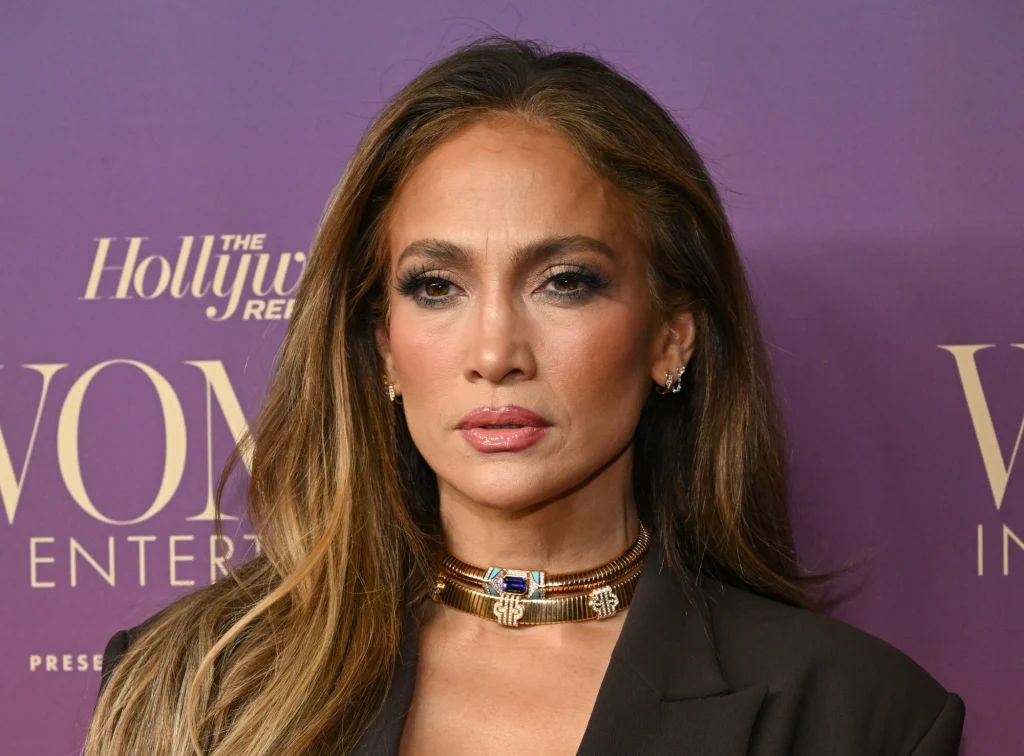 Kobieta o długich, rozpuszczonych włosach, formalnie ubrana, z wyrazistym makijażem, pozująca na tle fioletowej ścianki z napisem, nosząca elegancki naszyjnik.